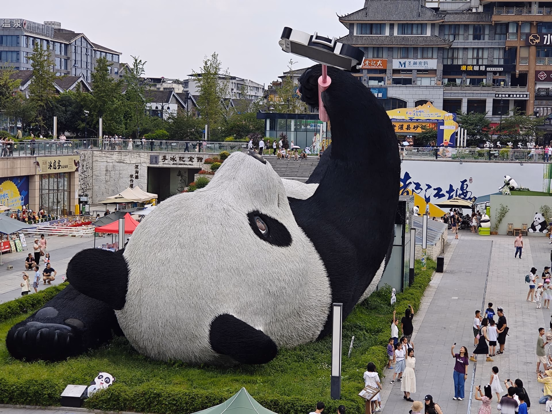 The image depicts a large panda sculpture lying on its back with one leg raised and people walking around it. (由 AI 生成标题)
