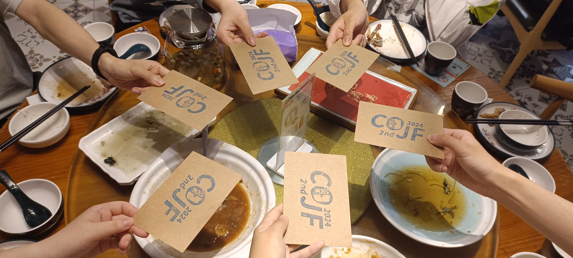 A group of people are holding up cards with the word "COU" on them, sitting around a table with various dishes and bowls.  (Captioned by AI)