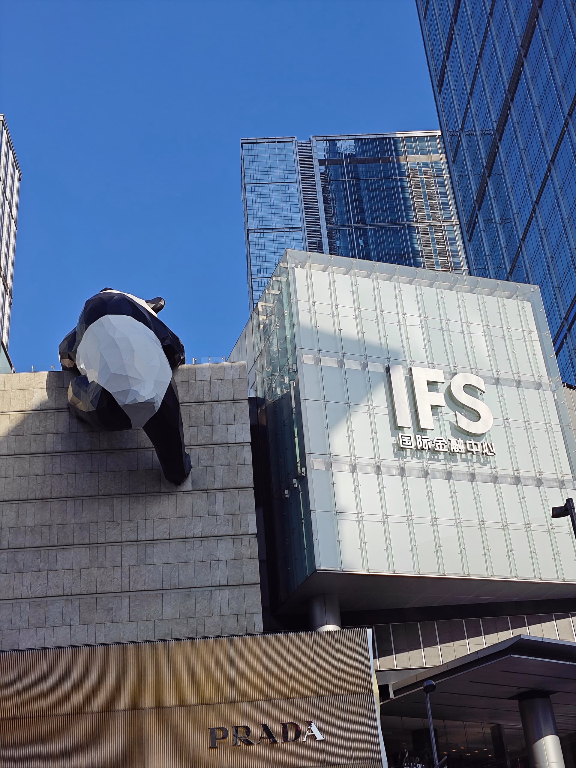 A large building with a panda sculpture on the side, featuring logos for IFS and Prada, set against a backdrop of other skyscrapers and a clear blue sky. (由 AI 生成标题)