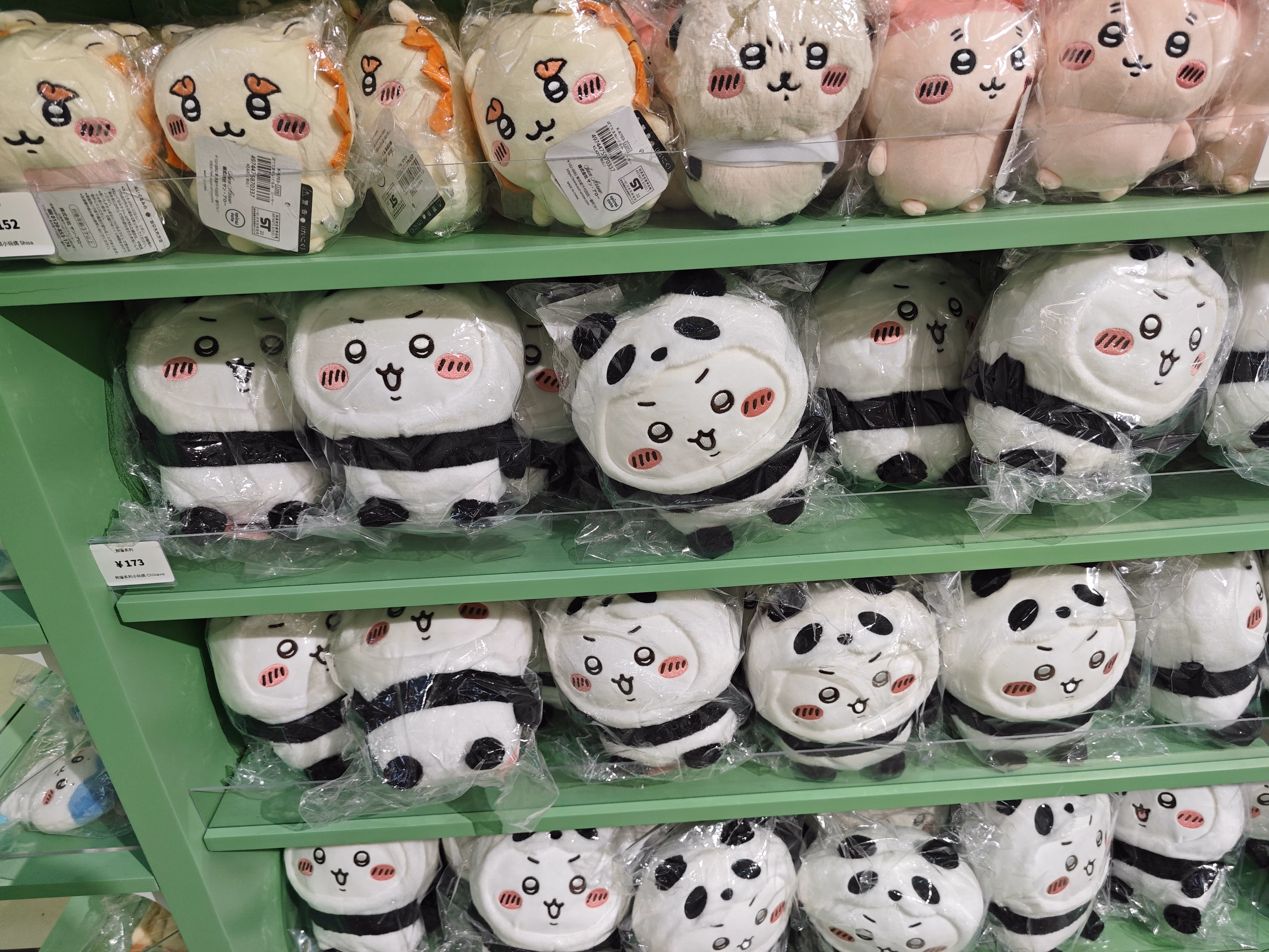 The image shows three green shelves stacked with plush toys individually wrapped in plastic and featuring cute, stylized faces. (由 AI 生成标题)