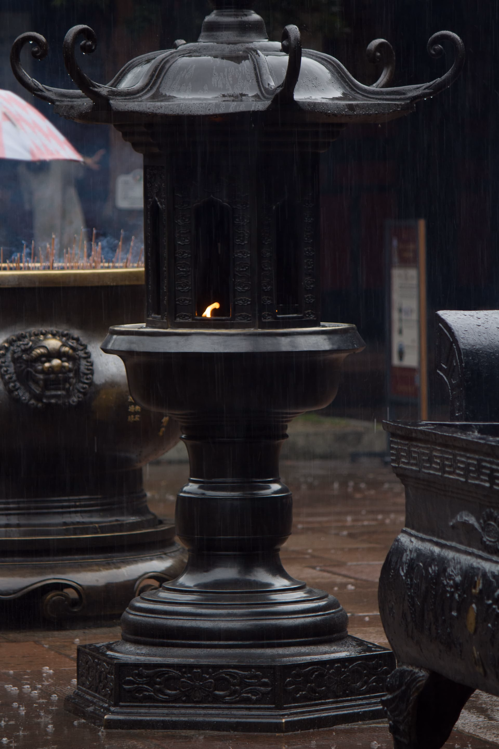 The image depicts a rainy scene featuring an ornate, black, pagoda-style lantern with a small flame burning inside, situated on a wet stone floor amidst other similar structures. (由 AI 生成标题)
